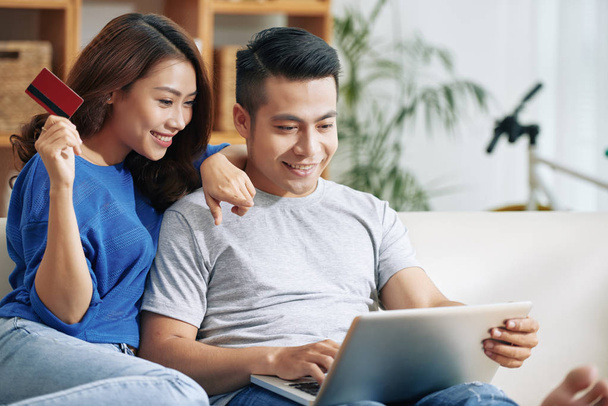 Pretty Asian woman and handsome man relaxing on couch with laptop and credit card making purchases online - Photo, Image