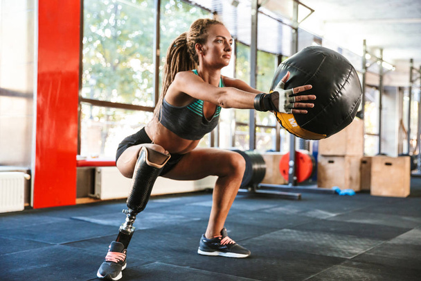 Portrait of european invalid woman with prosthesis in tracksuit doing sit-ups with fitness ball in gym - Foto, Imagem