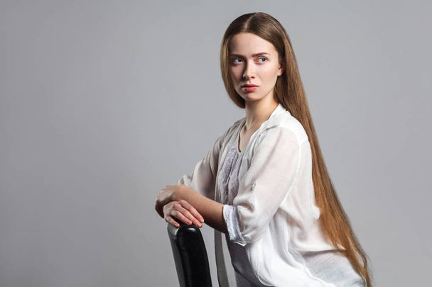 Portrait of thoughtful serious young model actor with long brown hair in white t-shirt sitting on black chair and looking away on grey background - Foto, afbeelding