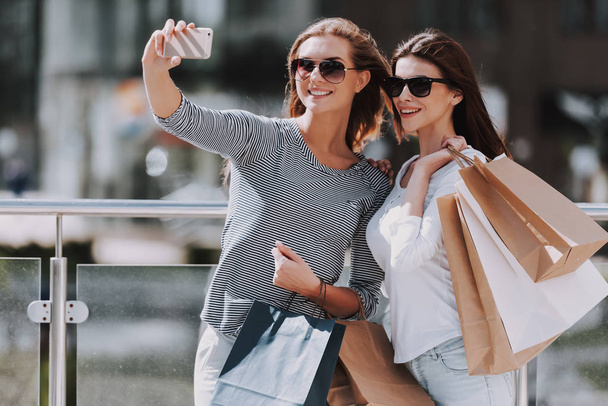 Female Friends Shopping at Mall and Taking Selfie. Sale, Consumerism and People Concept. Shopping and Tourism Concept. Girls Bags Walking Down the Street on Sunny Day after Good Shopping. - 写真・画像