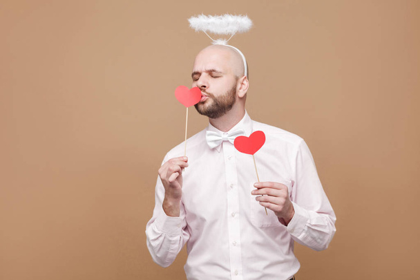 Happy handsome middle aged bald bearded angel in shirt and white halo decoration on head standing and kissing red heart sticker on brown background - Fotografie, Obrázek