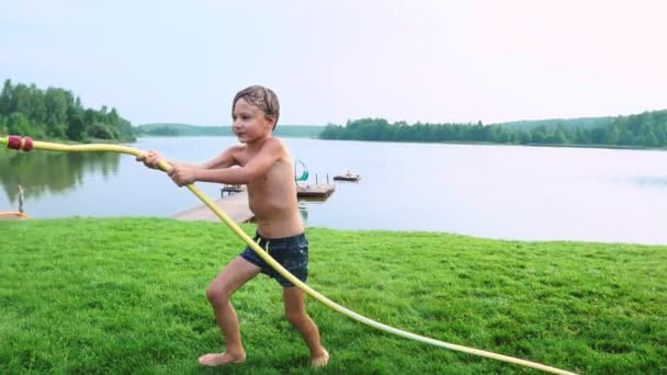 Junge in Badehose gießt Wasser auf seinen jüngeren Bruder, der sich im Park auf der Wiese am See vergnügt - Filmmaterial, Video