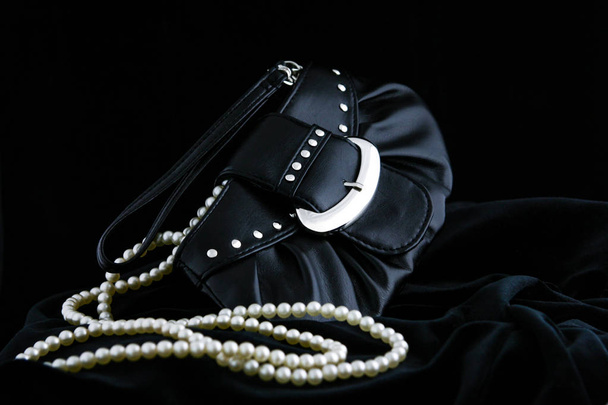 Элегантная, кожаная черная женская вечерняя сумка на черном велевете, изолированная на черном фоне. Жемчужные нити вываливаются из мешка. Концепция женского гламура и элегантности
 - Фото, изображение