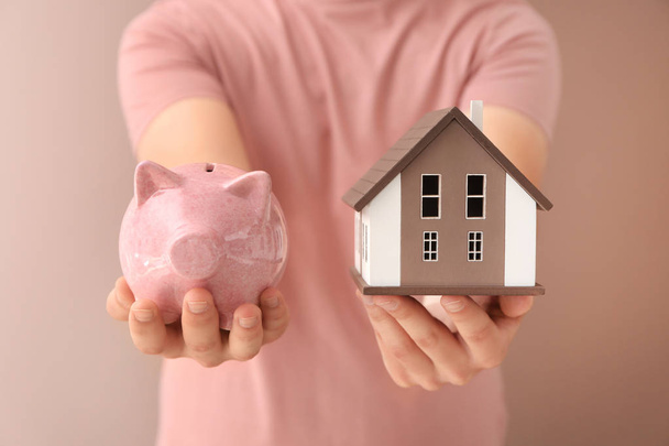 Man holding house model and piggy bank on color background. Mortgage concept - Photo, Image