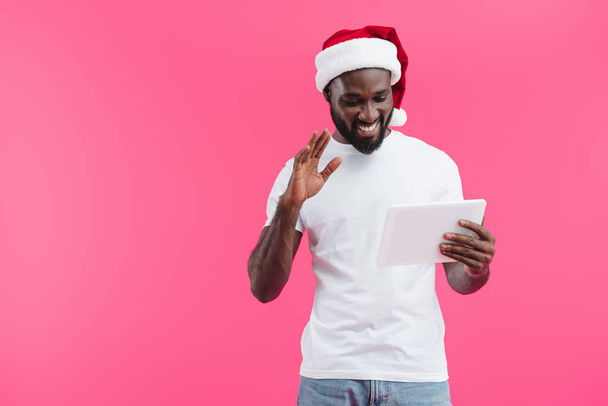 portrait of smiling african american man in santa claus hat with tablet gesturing isolated on pink  - Fotó, kép
