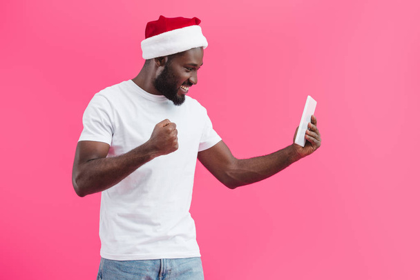 widok boczny uśmiechający się african american man w Czapka Mikołaja z tabletu na różowym tle  - Zdjęcie, obraz