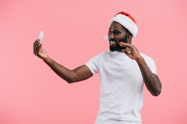 glimlachen van Afro-Amerikaanse man in Kerstman hoed met smartphone zwaaien geïsoleerd op roze  - Foto, afbeelding