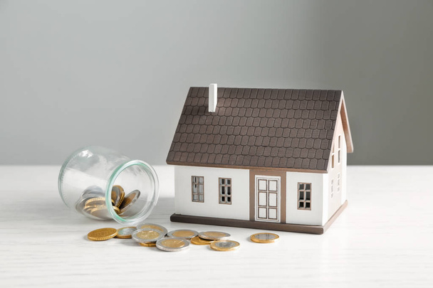 Hausmodell und Glas mit Münzen auf weißem Tisch. Hypothekenkonzept - Foto, Bild
