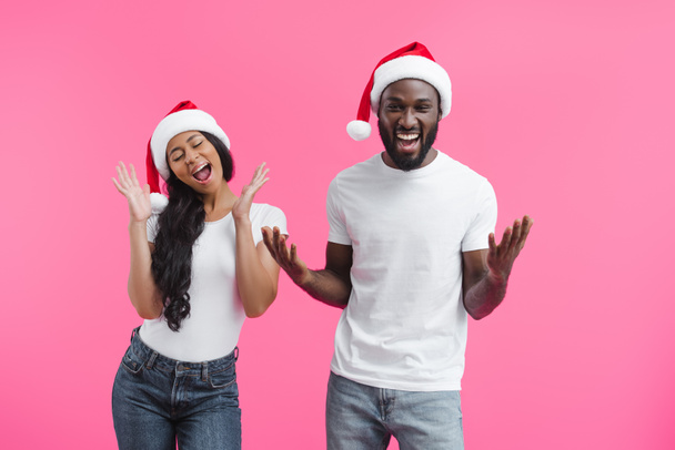excité couple afro-américain en chapeaux de Noël gestuelle par les mains isolées sur fond rose
  - Photo, image