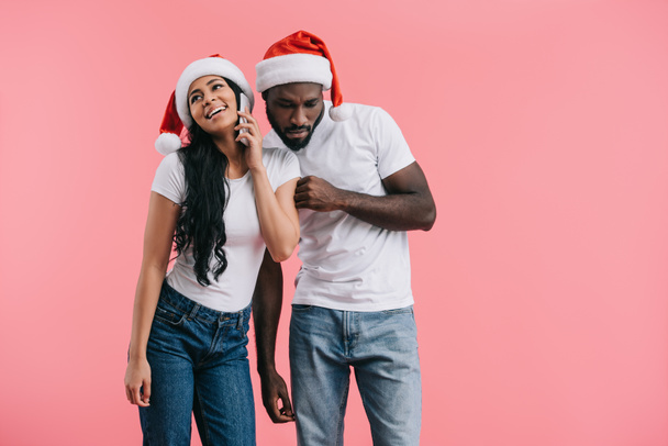 concentrated african american man in chrismas hat eavesdropping girlfriend talking on smartphone isolated on pink background  - Φωτογραφία, εικόνα