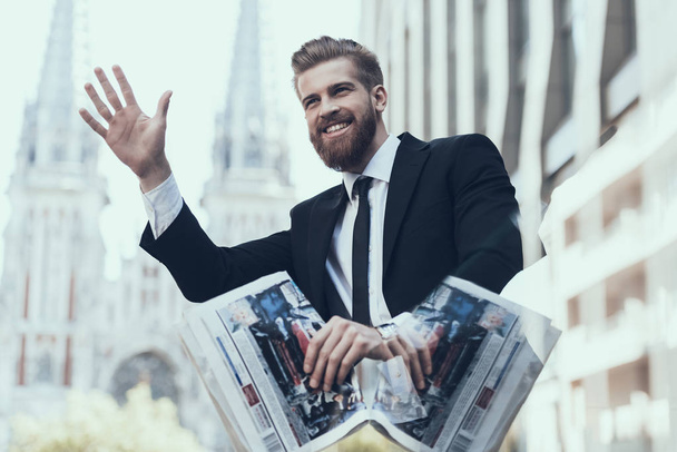 Smiling Handsome Businessman Meeting Somebody. Guy Handing Up. Man Waiting Somebody. Bearded Serious Man Wearing Suit and Glasses, Standing Outdoor and Reading Newspaper. Business Concept. - Zdjęcie, obraz