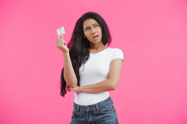 pembe arka plan izole prezervatif tutan şüpheci genç Afro-Amerikan kadın  - Fotoğraf, Görsel