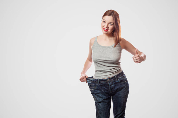 portrait of happy beautiful young woman with slim waist standing in big jeans and showing successful weight loss with thumb up on light gray background, diet concept - Foto, Imagen