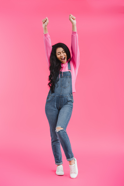 excited stylish young african american woman in denim overall with raised arms on pink background  - Foto, immagini