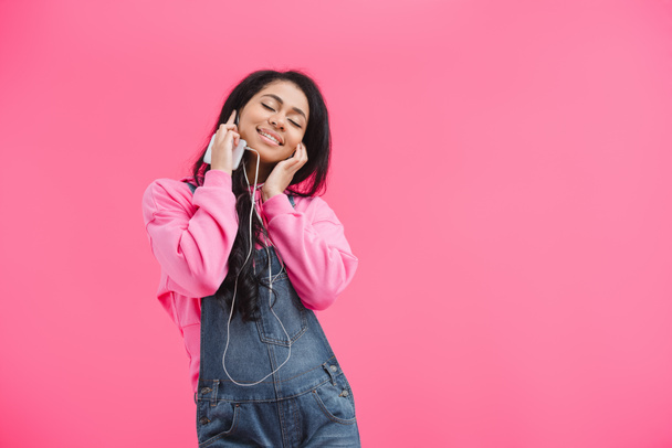 attractive african american woman with closed eyes in earphones listening music with smartphone isolated on pink background  - Fotografie, Obrázek