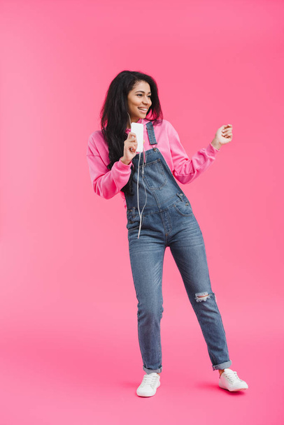 smiling african american woman in earphones listening music with smartphone dancing on pink background  - Foto, imagen