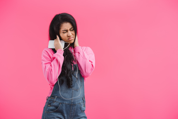 mujer afroamericana disgustada en auriculares escuchando música con teléfono inteligente aislado sobre fondo rosa
  - Foto, imagen