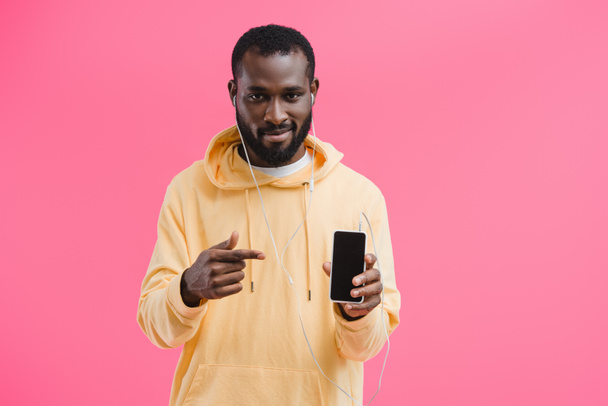 jovem afro-americano em fones de ouvido apontando para smartphone com tela em branco isolada em fundo rosa
 - Foto, Imagem