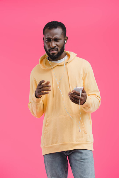 disappointed african american man in earphones listening music with smartphone isolated on pink background - Fotó, kép