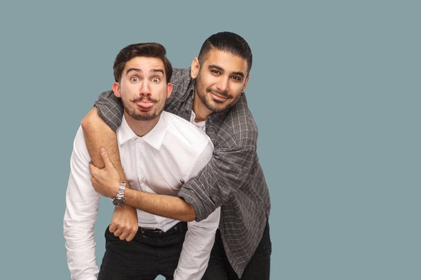 portrait of two handsome happy bearded friends enjoying and hugging while looking at camera with smile and funny face on blue background - Φωτογραφία, εικόνα