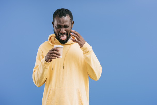 upset young african american man with grimace on face holding disposable coffee cup isolated on blue background - Photo, Image
