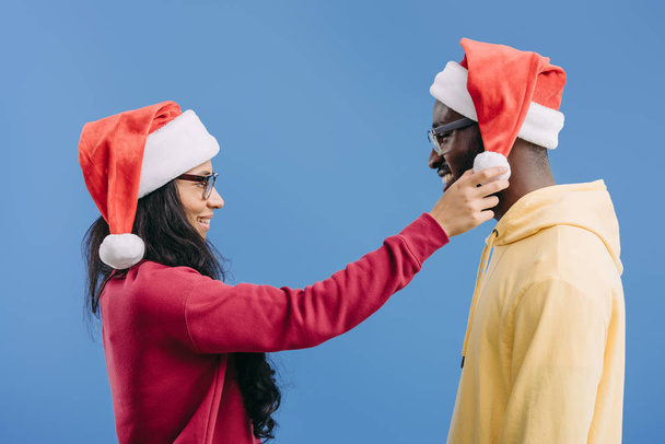 side view of african american woman adjusting christmas hat on boyfriend head isolated on blue background - Foto, immagini