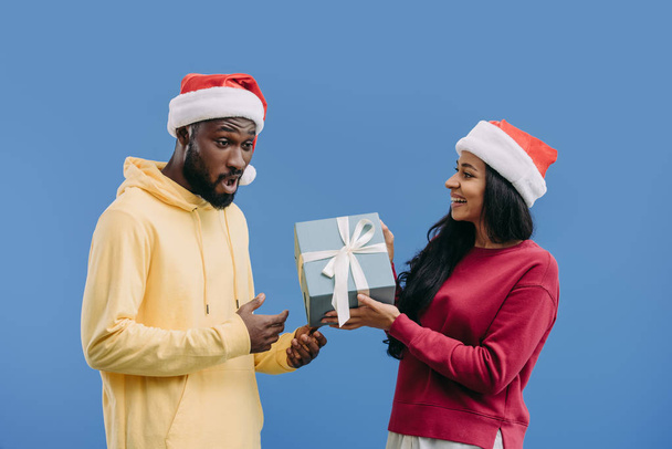 attractive african american woman in christmas hat giving gift box to shocked boyfriend isolated on blue background  - Foto, imagen