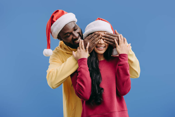 souriant homme afro-américain dans le chapeau de Noël fermer les yeux de la petite amie isolée sur fond bleu
 - Photo, image