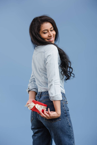 smiling attractive african american girl holding red gift box isolated on blue - Foto, Imagen
