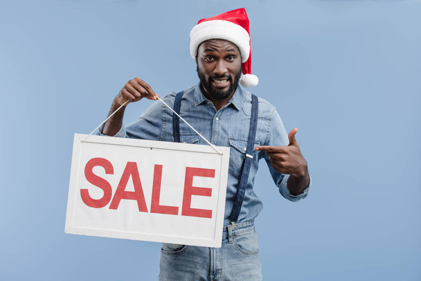 Grijnzende knappe Afro-Amerikaanse man in KERSTMUTS wijzen op verkoop teken geïsoleerd op blauw - Foto, afbeelding