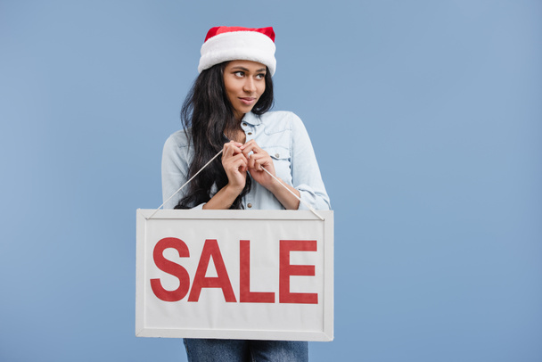 cheerful attractive african american girl in santa hat holding sale sign isolated on blue - Фото, изображение