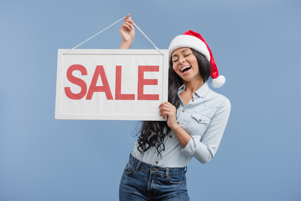 excited attractive african american girl in santa hat holding sale sign isolated on blue - Photo, Image
