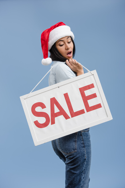 attraktive schockierte afrikanisch-amerikanische Mädchen in Weihnachtsmütze mit Verkaufsschild isoliert auf blau - Foto, Bild