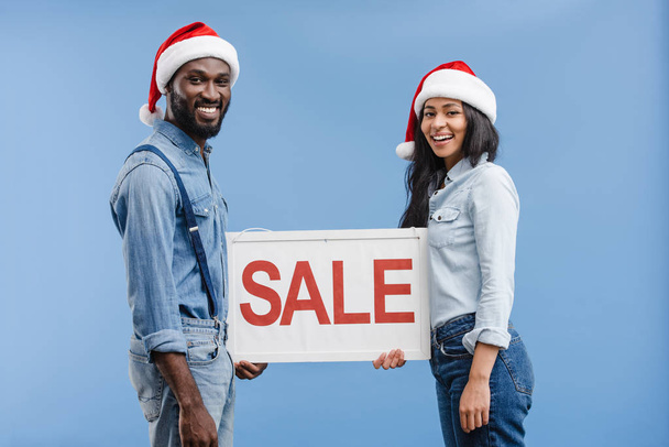 smiling african american couple in santa hats holding sale sign isolated on blue - Φωτογραφία, εικόνα