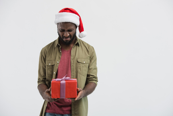 upset african american man in santa hat looking at present box isolated on white - 写真・画像