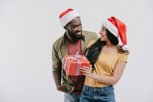 smiling african american couple in santa hats looking at each other and holding present isolated on white - Valokuva, kuva