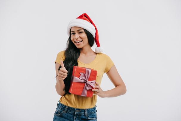 smiling african american girl in santa hat holding present and showing thumb up isolated on white - Zdjęcie, obraz