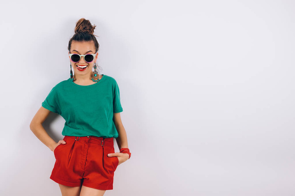 赤いショート パンツと白い壁の近くに立って、笑顔のグリーンの t シャツで身に着けているサングラスの美しい流行に敏感な女性. - 写真・画像