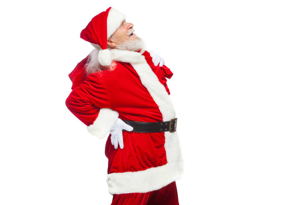 Christmas. Santa Claus is suffering from back pain and holds a red bag with gifts on his back. Isolated on white background. - Photo, image