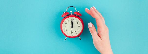 Creative flat lay concept top view of woman hand holding the red vintage alarm clock on bright blue turquoise color paper background with copy space in minimal style, long wide banner - Photo, Image