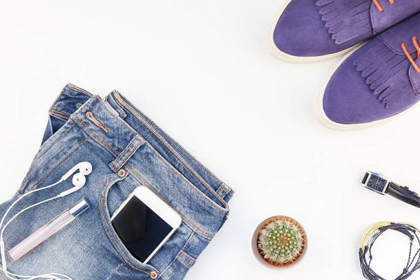 Flat lay of suede shoes, blue jeans, bracelets cactus and smartphone with headphones on white background with copy space. Overhead view of woman casual outfit. Trendy hipster look top view - Fotografie, Obrázek