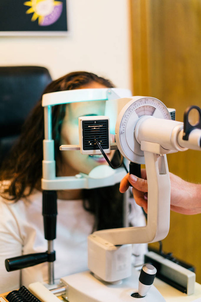 Oftalmólogo en sala de examen con mujer joven sentada en silla mirando a la máquina de prueba ocular
 - Foto, Imagen
