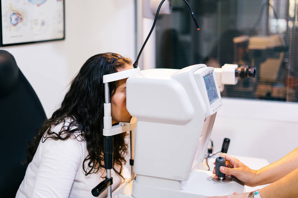Ophthalmologist In Exam Room With Young Woman Sitting In Chair Looking Into Eye Test Machine - Фото, изображение