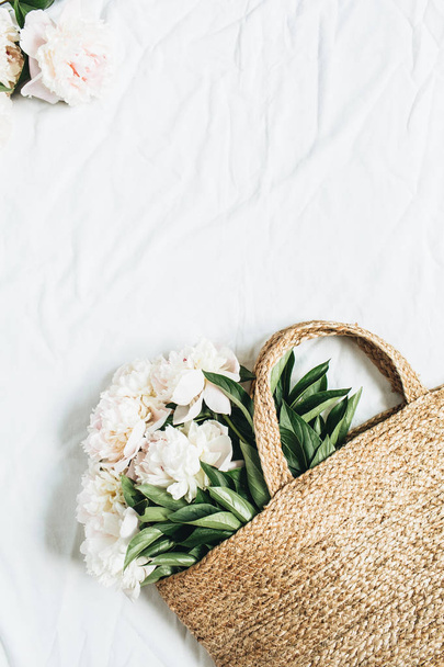 Straw bag with white peony flowers on white background. Floral flat lay, top view. - Foto, Imagen