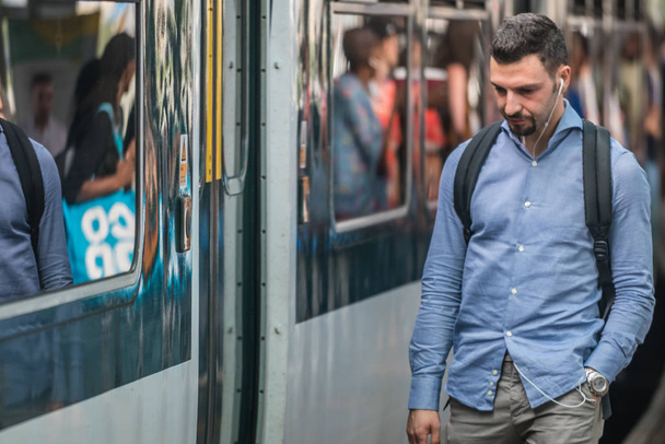Λονδίνο, Αγγλία - Ιουλίου 2018: Ο άνθρωπος ακούει τη μουσική στα ακουστικά κατά την αναμονή για τις υπέργειες τρένο που φθάνουν στην πλατφόρμα, Ηνωμένο Βασίλειο - Φωτογραφία, εικόνα
