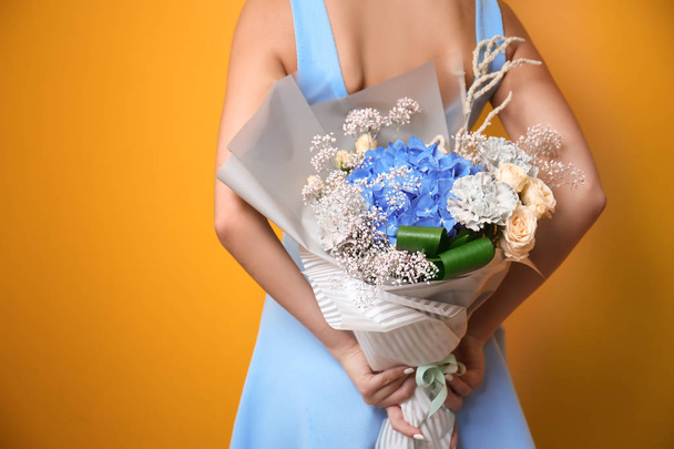 Young woman holding bouquet of beautiful flowers behind her back on color background - Foto, Imagen