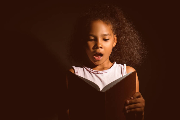 Surprised African-American girl reading book on dark background - Photo, Image
