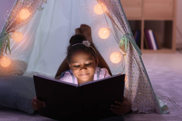 Little African-American girl reading bedtime story in hovel - Foto, Imagen