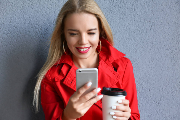 Mooie jonge vrouw met mobiele telefoon en koffie in de cup in de buurt van kleur muur - Foto, afbeelding