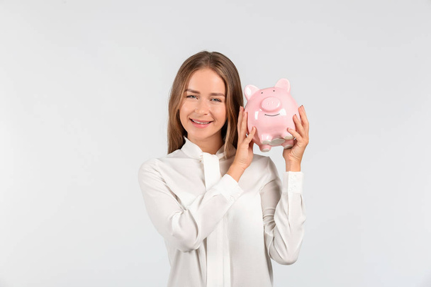 Happy young woman with piggy bank on light background - 写真・画像